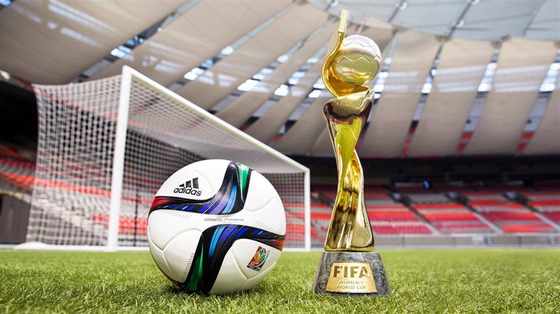 Soccer ball and World Cup trophy sit on a the grass of a stadium.
