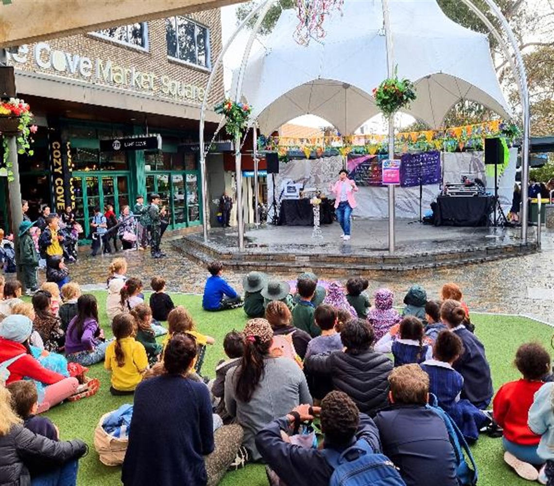 Children sitting on the Plaza green while a performer takes to the stage